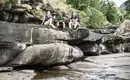 Mittagspause auf unserer Wanderung in den Drakensberge Drei Personen sitzen auf Felsen in den Drakensbergen, umgeben von einem Wasserlauf, mit Aussicht auf die Berglandschaft.