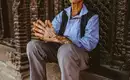 Weiser Nepalesischer Mann in Bhaktapur Ein älterer nepalesischer Mann mit Brille und traditionellem Hut sitzt vor einem kunstvollen Tempel in Bhaktapur.