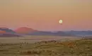 Mondaufgang über Namibias Wüstenlandschaft Vollmond über der weiten Wüstenlandschaft Namibias bei Sonnenuntergang, mit Bergen im Hintergrund.