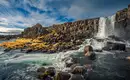 Wasserfall und Felsen entlang der Küste Islands Ein beeindruckender Wasserfall auf Island mit großen Felsen und fließendem Wasser im Vordergrund.