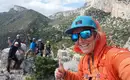 Eine Gruppe von Wanderern posiert auf einem Felsvorsprung in den Bergen. Eine Person im Vordergrund macht ein Selfie mit Daumen hoch. Alle sind in Outdoor-Bekleidung und tragen Kletterhelme.