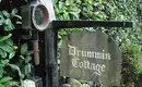 Historisches Eingangsschild zum Drummin Cottage Schild mit der Aufschrift 'Drummin Cottage' an einem hölzernen Torpfosten, umgeben von grüner Vegetation.