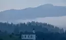 Morgenstimmung in den Alpen Eine Kirche mit Zwiebelturm vor nebeligen, bewaldeten Alpenlandschaft.