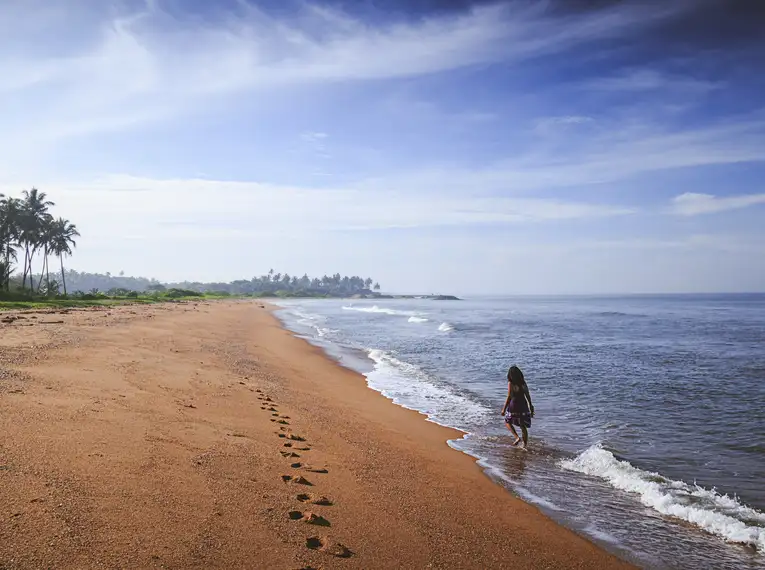 Person geht am ruhigen Sandstrand in Sri Lanka entlang, mit Palmen im Hintergrund.