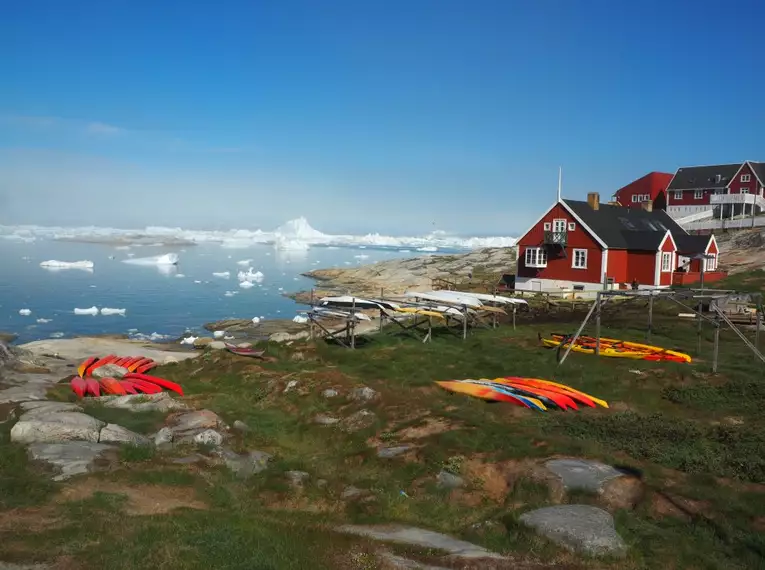 Bunte Kajaks vor grönländischem Dorf Rote Häuser und bunte Kajaks am Meer, Eisberge im Hintergrund in Grönland.