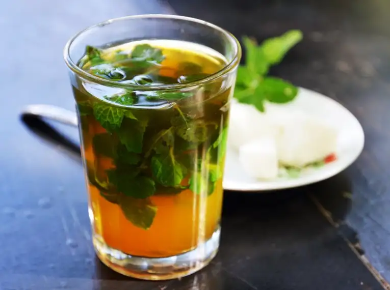 Marokkanischer Pfefferminztee - so lecker! Belebender Pfefferminztee im Glas mit frischen Minzblättern und Zuckerwürfeln auf einem Teller daneben.