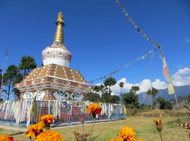 Bunte Stupa im Himalaya mit Gebetsfahnen und Blumen im Vordergrund, blauer Himmel im Hintergrund.