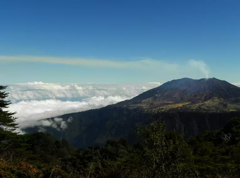 Rauchender Vulkan in Costa Rica mit bewaldeter Landschaft im Vordergrund und Wolken am Horizont.