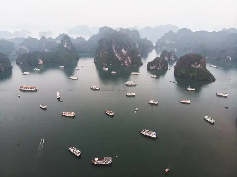 Luftaufnahme vieler Boote in der Halong-Bucht, umgeben von markanten Karstfelsen und trübem Himmel.