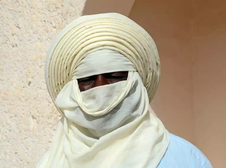 Eine Person in traditioneller Tuareg-Kleidung mit hellem Gesichtstuch vor einer Steinwand. Sahara-Reisen.