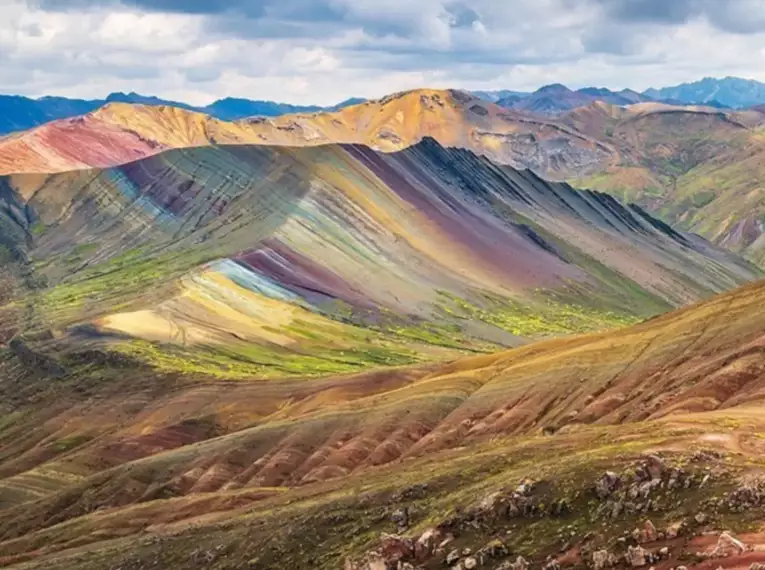 Bunt gestreifte Mineralien formen die Regenbogenberge von Palccoyo in Peru, umgeben von einer weiten, hügeligen Landschaft.