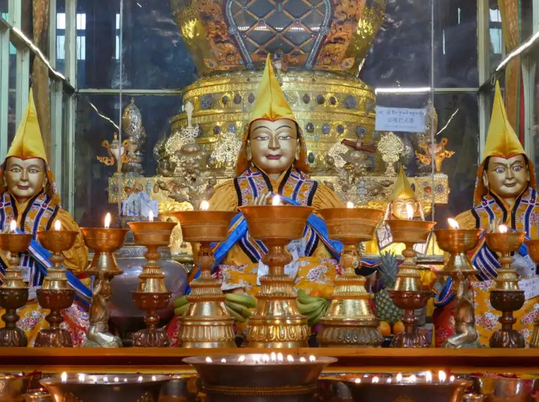 Mehrere bunt gekleidete buddhistische Statuen stehen in einem tibetischen Tempel mit Opfergaben und leuchtenden Kerzen davor.