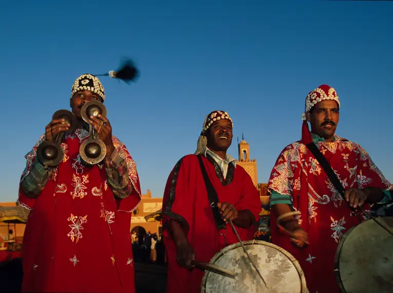 Drei Musiker in traditionellen marokkanischen Gewändern spielen Instrumente unter blauem Himmel.