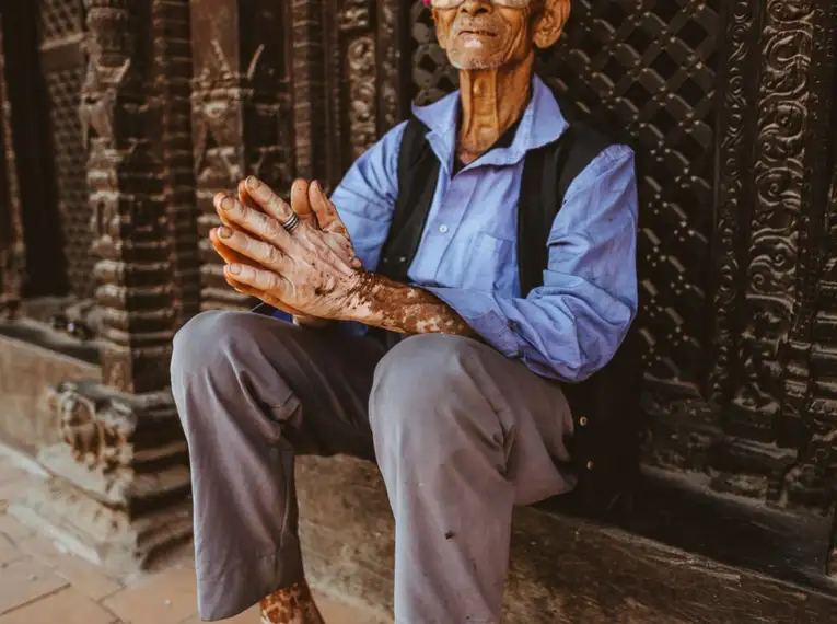 Ein älterer nepalesischer Mann mit Brille und traditionellem Hut sitzt vor einem kunstvollen Tempel in Bhaktapur.
