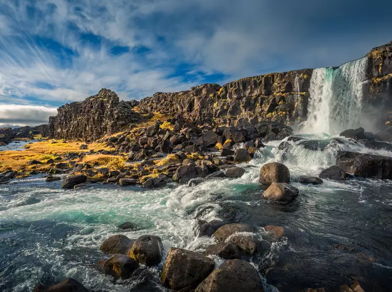 Wasserfall und Felsen entlang der Küste Islands Ein beeindruckender Wasserfall auf Island mit großen Felsen und fließendem Wasser im Vordergrund.