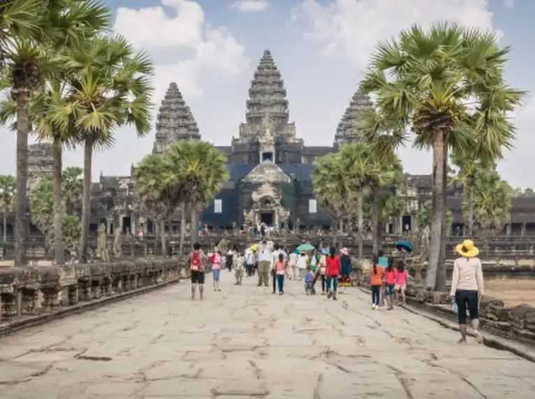 Besucher betreten Angkor Wat vor Palmen und blauem Himmel.