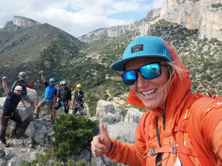 Eine Gruppe von Wanderern posiert auf einem Felsvorsprung in den Bergen. Eine Person im Vordergrund macht ein Selfie mit Daumen hoch. Alle sind in Outdoor-Bekleidung und tragen Kletterhelme.