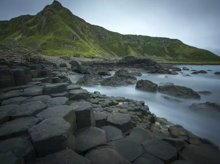 Basaltsäulen am Giant's Causeway Hexagonale Basaltsäulen am Ufer mit grünem Hügel im Hintergrund