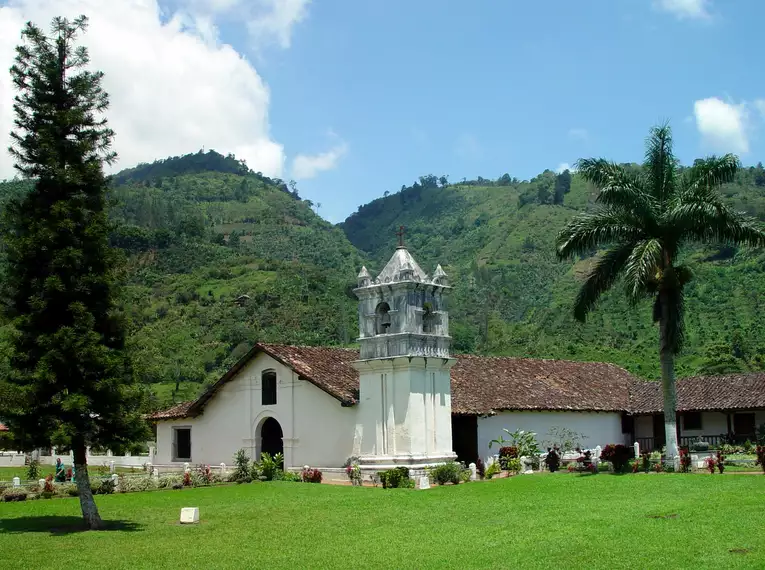 Eine kleine, weiß getünchte Kirche vor grünen, bewaldeten Hügeln und Palmen.