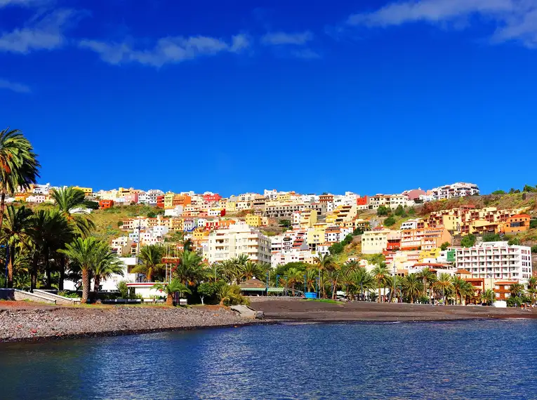 Bunte Gebäude vor einem blauen Himmel an der Küste von La Gomera, umgeben von Palmen.