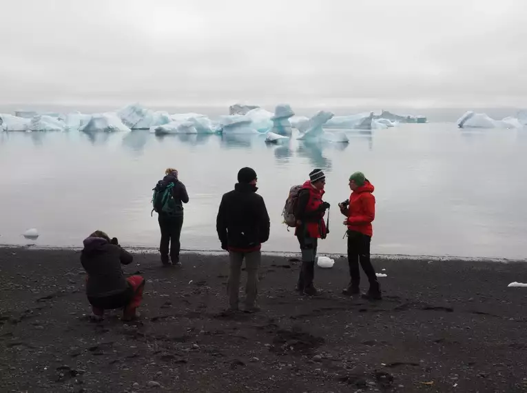 Gruppe vor eisiger Küstenlandschaft Reisende stehen am Strand mit mehreren Eisbergen im Hintergrund in Grönland.