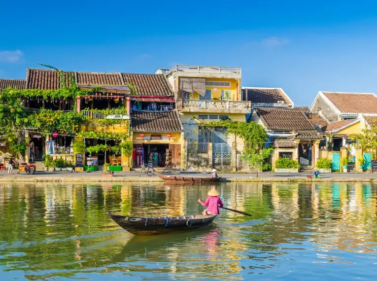 Fluss mit traditioneller Holzboot und bunten Häusern in Hoi An bei Sonnenschein.
