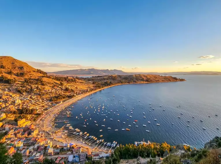 Ein Panoramablick auf eine Bucht des Titicacasees mit Bergen und Booten im goldenen Sonnenuntergang.